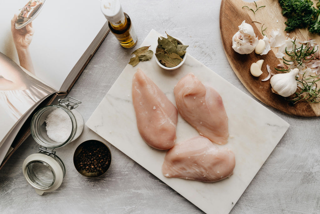 Raw chicken breasts on a marble slab with herbs and spices on a light wooden surface.